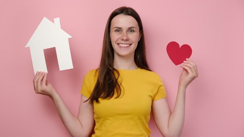 Friendly attractive young female holding white paper house with small red heart, happy looking at camera, wears t-shirt, model over pink studio background. Charity, real estate and family home concept
