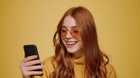 Cheerful girl watching video on phone in studio. Closeup happy woman looking smartphone screen on orange background. Portrait of joyful female person reading message on cellphone indoors.