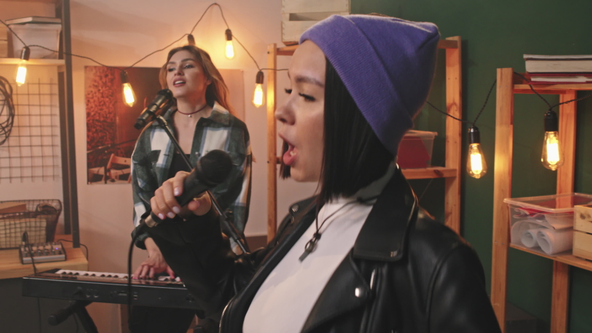 Handheld medium slowmo of young cool all-female music band playing rock music in retro-style studio. Attractive vocalist in stylish beanie hat and leather jacket singing emotionally in microphone Royalty-Free Stock Footage #1072343573