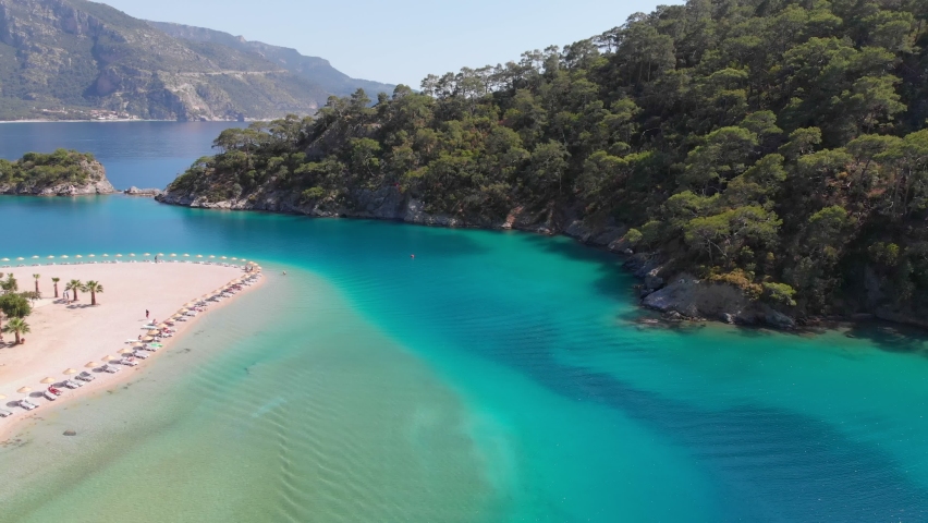People sunbathe on the sandy beach on the shores of the azure bay. Aerial view of azure water in mediterranean sea in bay near Oludeniz, Turkey. Boat sails through the purest turquoise water | Shutterstock HD Video #1072344638