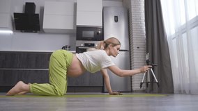 A young pregnant blogger records online video lessons for pregnant women on her smartphone, with a show of yoga exercises, a conversation with subscribers sitting at home on a fitness mat