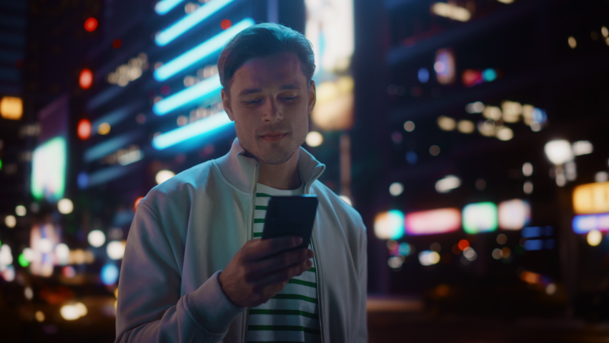 Portrait of Man Using Smartphone Walking Through Night City Street Full of Neon Light. Smiling Stylish Man Using Mobile Phone, Social Media, Online Shopping, Texting on Dating App. Medium Shot Royalty-Free Stock Footage #1072347206