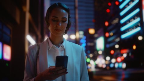 Beautiful Young Woman Using Smartphone Walking Through Night City Street Full of Neon Light. Portrait of Gorgeous Smiling Female Using Mobile Phone. Tracking Medium Close-up Shot