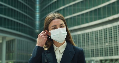 Cinematic close up  of young business woman taking off medical protective mask and smiling in camera on background of skyscrapers. Concept of corporate, protection, virus transmission, safety, covid-19