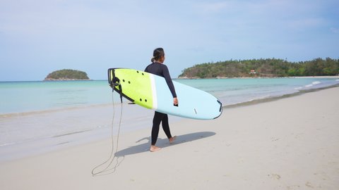 Confidence Asian woman in wetsuit holding surfboard and walking on tropical beach at summer sunny day. Healthy sportswoman enjoy outdoor active lifestyle play extreme sport surfing on summer vacation