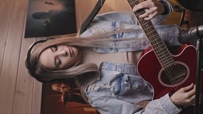 Vertical medium slowmo portrait of attractive young female guitarist in vintage oversize blue denim jacket looking at camera while playing acoustic guitar in old-school music studio Royalty-Free Stock Footage #1072356887