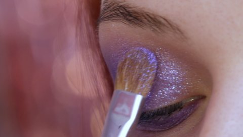 Beautiful shiny violet-lilac shadows are applied with a special brush on the eyelid of the model. The process of bright evening make-up close up view