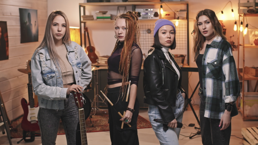 Medium portrait of four cool young women in grunge fancy clothes looking at camera standing in retro-style garage turned into music studio Royalty-Free Stock Footage #1072357982