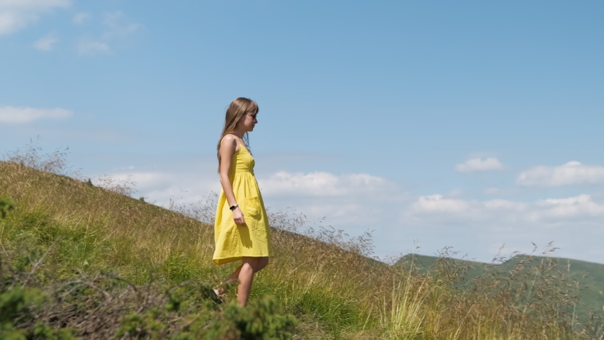 Young happy woman in yellow dress walking on hillside meadow on a windy day in summer mountains.
