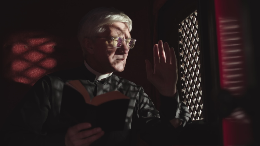 Medium shot of old Caucasian priest wearing black robe, white collar and eyeglasses sitting in confessional with Holy Bible in hands and listening to confession | Shutterstock HD Video #1072360322
