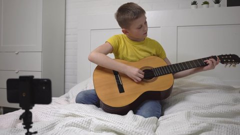 child blogger plays the guitar boy is sitting on sofa in front of phone and playing musical instrument video blogger Online guitar lessons Communication over Internet Learning coronavirus pandemic