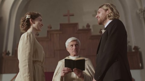 Low angle view of young Caucasian man and his beautiful bride standing at altar in Christian church and pronouncing vows during wedding ceremony while old priest reading Bible