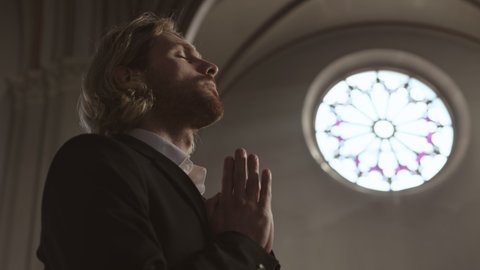 Low angle view of young Caucasian red-haired man wearing jacket and white shirt standing in Christian church, crossing himself and then going away