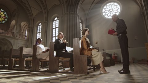 Wide shot of African-American pastor wearing black trousers, shirt and white collar standing in Lutheran church with Bible in hands and talking to one of the parishioners