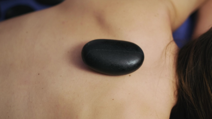 Hot Stone Therapy is a form of massage using heated stones. masseuse is putting stones on woman's back in slow motion. 4K.