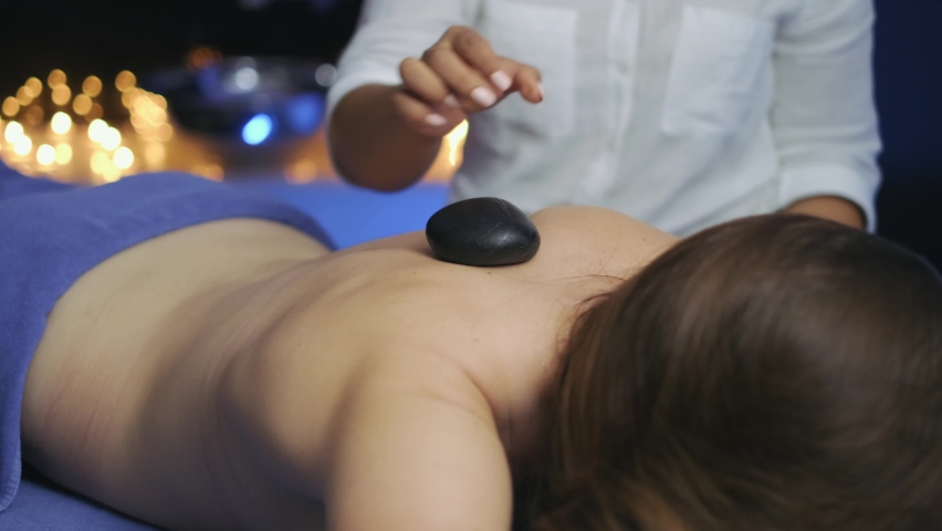 In the massage room, the masseuse is putting hot stones on the woman's back. Burning candle lights in the background. slow motion. 4K.