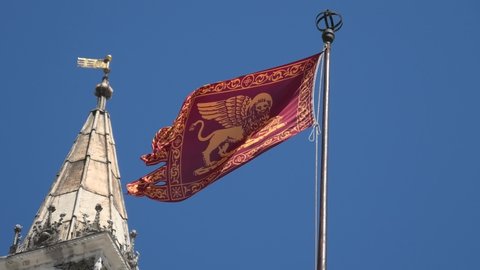 Close-up of San Marco flag, Saint Mark in Italy waving on sky background with Italian Saint Mark Basilica. Red flag of Venice city and Veneto region, with a golden-winged lion.