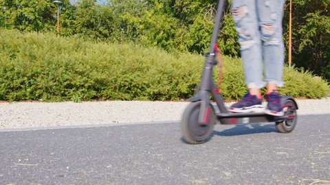 Legs of a female person in blue jeans and sneakers moving on an electric scooter. A woman riding a kick scooter in the park