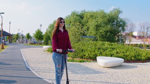 A woman is riding an electric scooter in the park on a summer day. A young female wearing red plaid shirt and blue jeans is spending a weekend outdoors