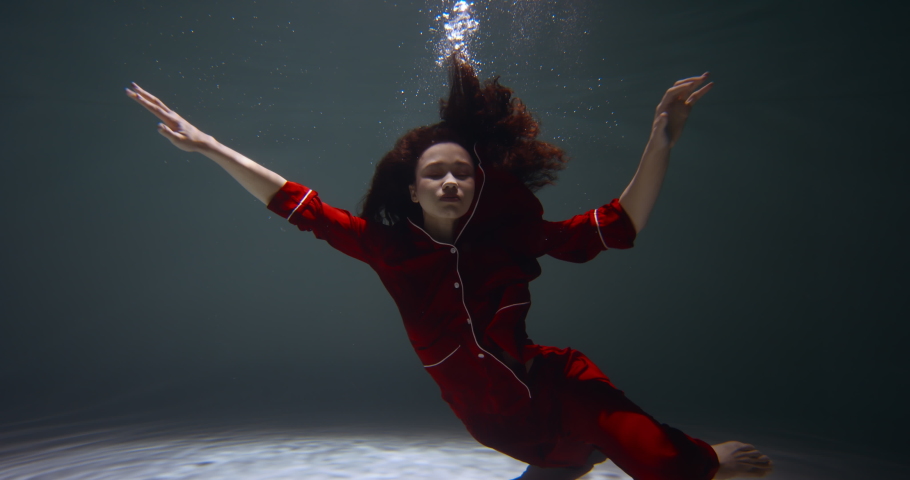 Ethereal cinematic underwater portrait of young beautiful woman in red pyjamas looking at camera, arms open slow motion.