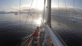 A video taken from the side of a sailboat going through the ice filled ocean of Antarctica. The sun is setting over the horizon and the orange light reflects off of the water and icebergs.