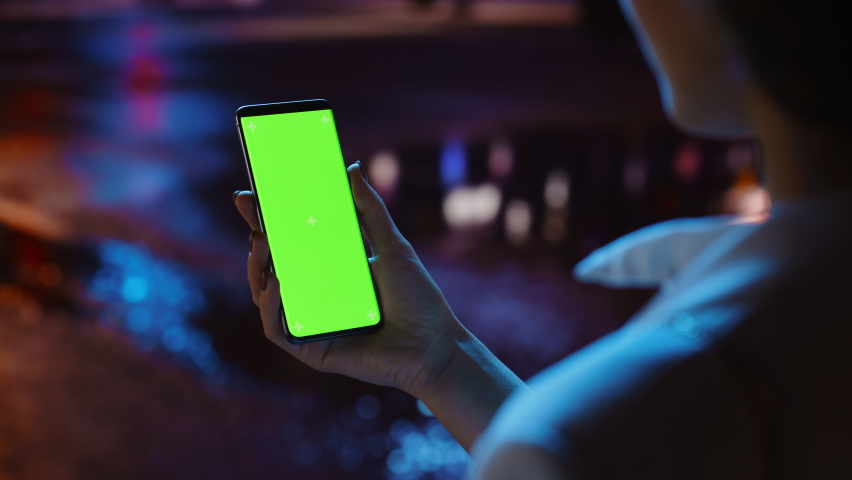Beautiful Woman using Chroma Key Smartphone while Walking Through Night City Street Full of Neon Light. Female Using Green Screen Mobile Phone. Over the Shoulder Static Medium Close-up Shot Royalty-Free Stock Footage #1072389917