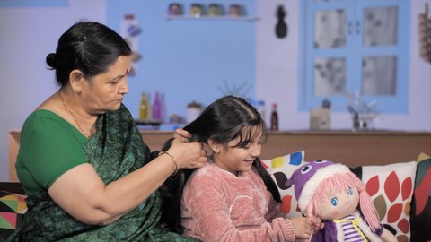 Loving grand mom tying a little girl's braids while she is playing with her doll - Indian family. Medium shot of a loving elderly woman taking care of her grandchild's hair - relationship and bonding