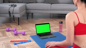 Green screen mock up chroma key monitor laptop: Fit woman in sportswear sits on yoga mat looks computer display, tells listen fitness trainer online video call webcam chat, distance sport course