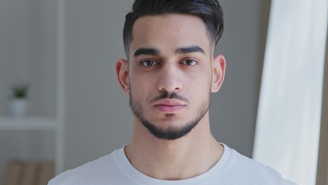 Portrait of serious handsome young adult arabic man with beard in white t-shirt stands posing indoors at home looking at camera. Close-up sad hispanic male ethnic guy brunette freelancer husband model
