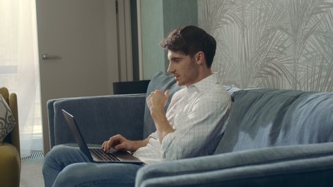 Side view handsome guy using laptop in modern interior space. Smiling man receiving message on computer indoors. Young businessman chatting on netbook on couch in hotel room.