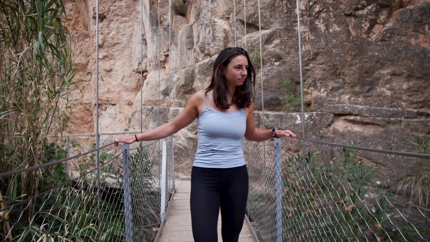 Young woman walking down a bridge and overlooking natural rock canyon in Chulilla, Spain. Mid angle, traveling movement, 4K.