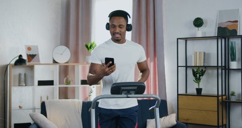 Home workout concept where handsome smiling satisfied active young black-skinned guy training on running track at home and enjoying favourite songs in headphones