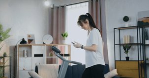 Pretty positive active slim young brunette in training clothes enjoying pleasant video chat with friend during workouting on treadmill at home,slow motion