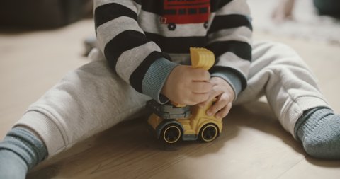 View of baby boy playing excavator on the wooden floor. Mothers hand shows him how to dig. Room filled with morning sunlight. Tiny fingers with toys.