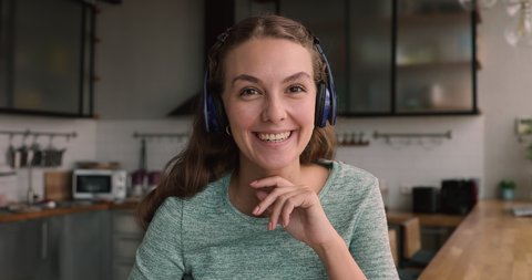 Head shot portrait young woman sit in kitchen wear wireless headphones wave hand having conversation by videocall, participate in job interview virtual meeting, talk to family living abroad concept
