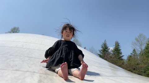 Asian little toddler girl playing in the park playground. Sliding down a huge white trampoline. Hair is floating and spreading due to static electricity. 3 years old child in black dress. Outdoor