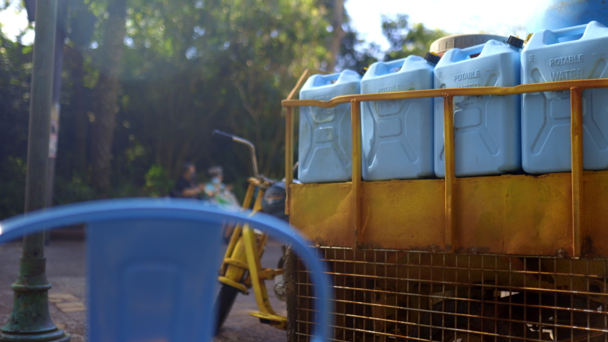 Colorful vintage yellow cargo motorcycle and blue chair with trees and bright sunlight as background. Tall wall of green leaves behind three-wheel motorbike with plastic gas tanks. Rustic outdoors