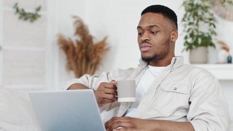 Portrait afro man african black male freelancer student mixed race ethnic guy sitting on couch at home cozy living room holding cup with tea coffee drink uses laptop listening to music online singing