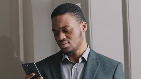 Frustrated African American guy look at cell phone screen reads bad news in mobile message concept, feels shocked. Worried businessman lost money thinking over sudden problems after reading sms