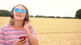 Slow motion video clip of pretty blonde girl teenager young woman wearing a striped t- shirt and blue sunglasses listening to music on her cell phone and wireless headphones 