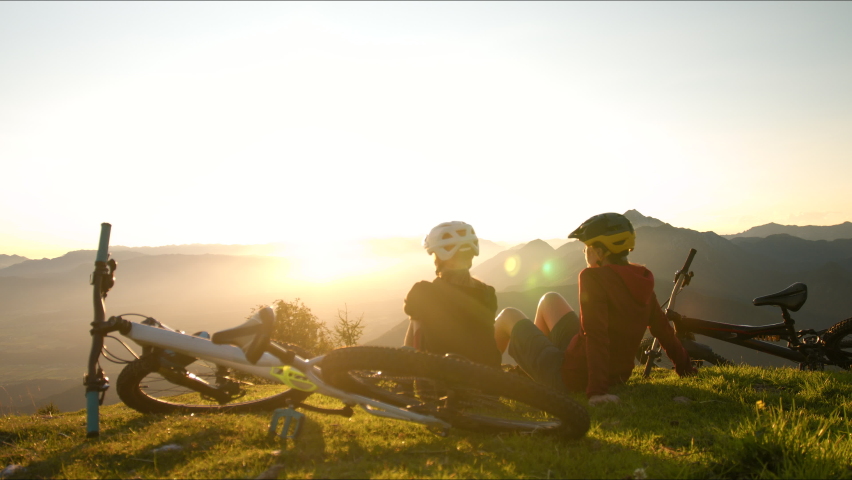 PAN right shot of a two female silhouettes, mother and daughter cyclists talking and sitting on the meadow at the top of the hill, the bikes are laid on the grass