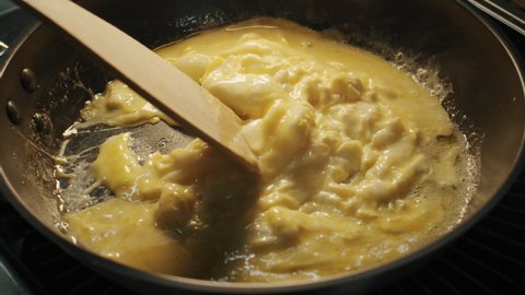 A semi-cooked tender scramble eggs are stirred with a wooden spatula in a hot frying pan
