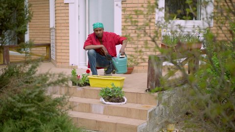 Extreme wide shot of male gardener watering ground in flower pot outdoors. Portrait of young African American man taking care of plants on porch in garden. Floristics and landscaping