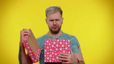 Handsome stylish tourist man unwrapping gift and expressing disappointment, dislike to bad present, man dissatisfied with birthday surprise. Young adult guy indoor isolated on yellow studio background