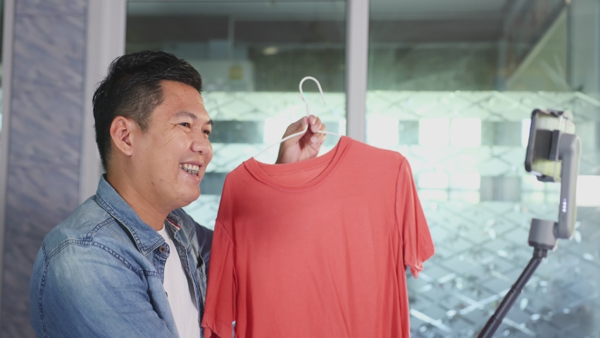 Entrepreneur of Asian young man live streaming video online for selling her dress and clothes to the customer in platform or application online, a hobby concept during a COVID-19 quarantine period.