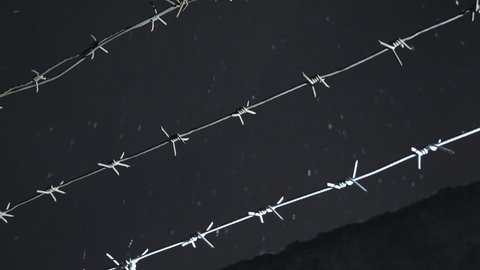 prison barbed wire over concrete fence against cloudy sky at cold dark winter night during snowfall. incarceration, jail, restricted area, closed zone, security concept