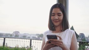 Happy young Asian woman blogger tourist with casual style hold smartphone look with smile joyful with beautiful sunset on river view near cafe in city town. Lifestyle tourist travel holiday concept.