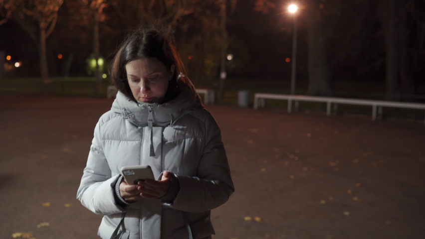 Attractive adult woman in warm coat holding cell phone in the street at cold night, brunette lady with pretty face typing on mobile phone screen on an empty autumn street in evening.