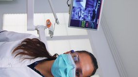 Vertical video: Patient pov to dentist talking about mouth hygine showing x-ray on monitor. Stomatology specialist wearing protective face mask and gloves pointing at teeth radiography in