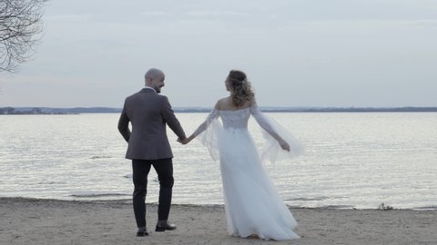 Back view of Happy sincere beautiful young couple bride and groom walking on a beach coast near sea, laughing, enjoying time together on sunset, awesome landscape with white swans